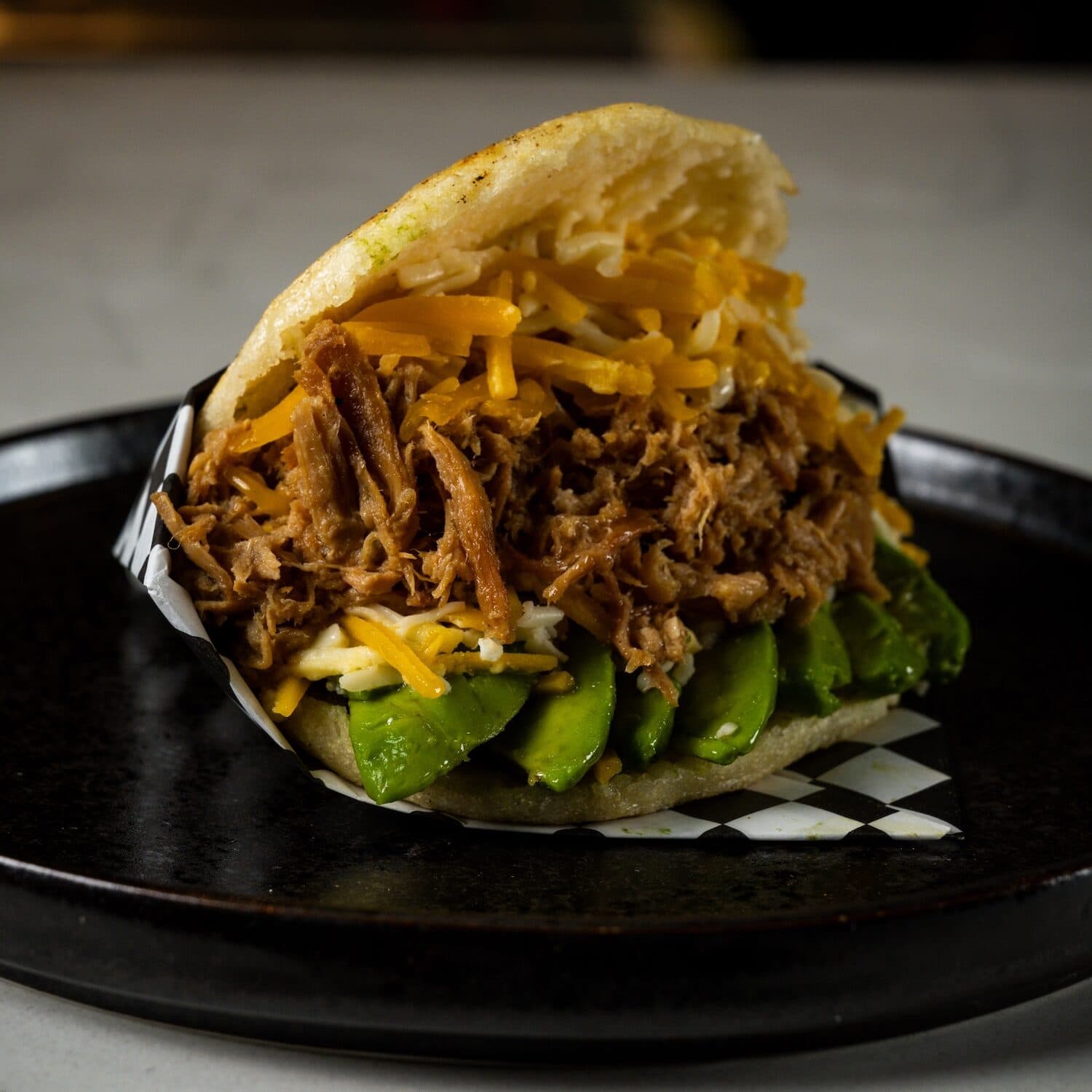 Meat based arepa with grated cheese and avocado slices on a black plate