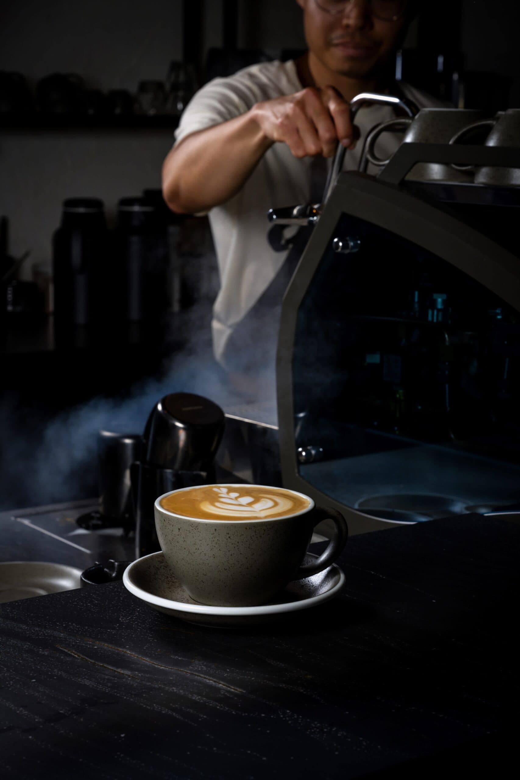 Latte in a stoneware bowl in the foreground and barista steaming milk in the background