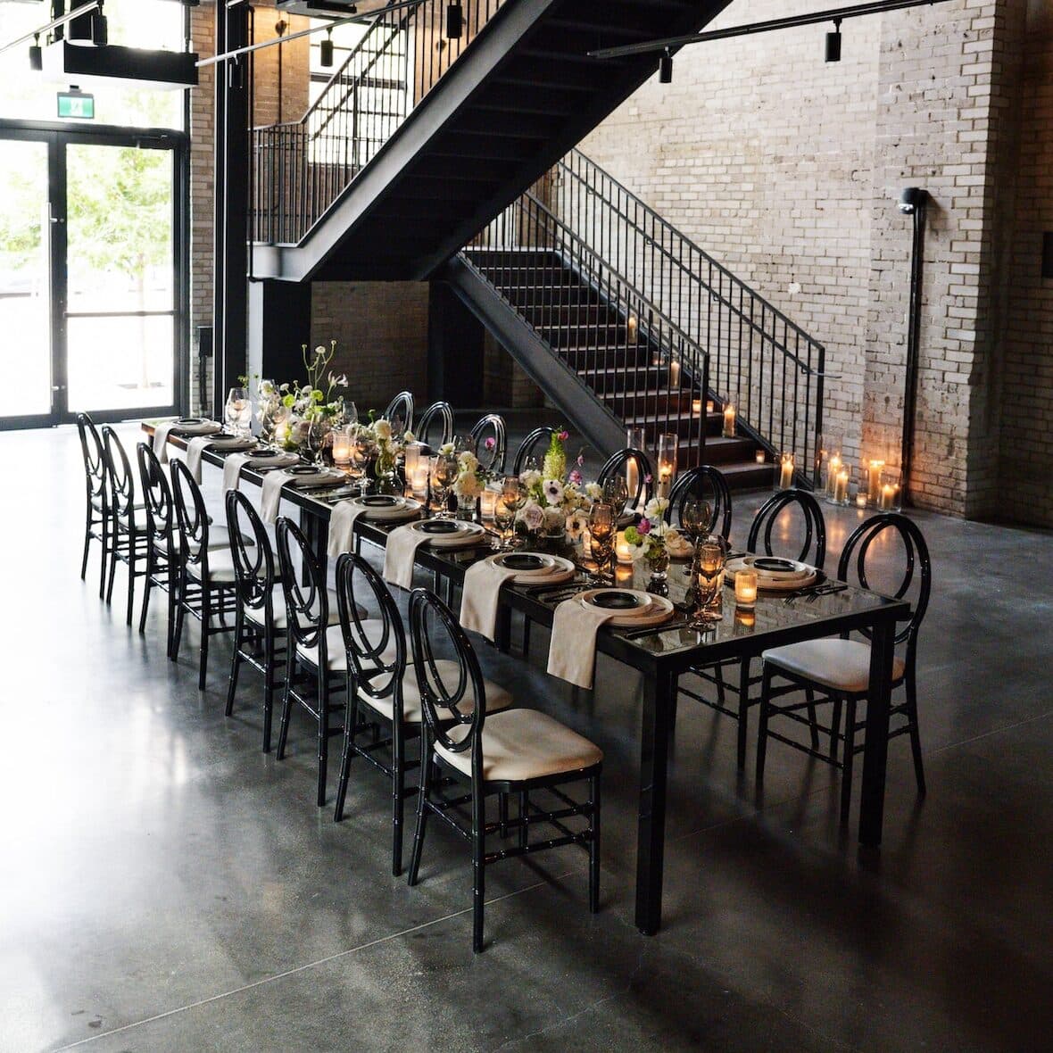 Seated dinner for 16 set in the middle of the Reception Room of the Event Hall. The stairs leading to the West Hall are in the background and contrast against the white brick walls.