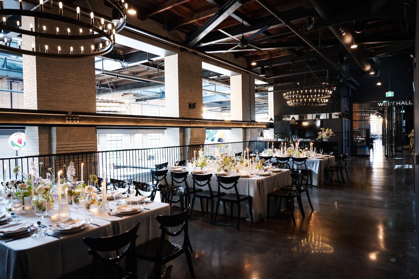 Seated dinner set up in the east hall showing square tables draped in light gray linens with organic flower arrangements and tapered candles in the middle.