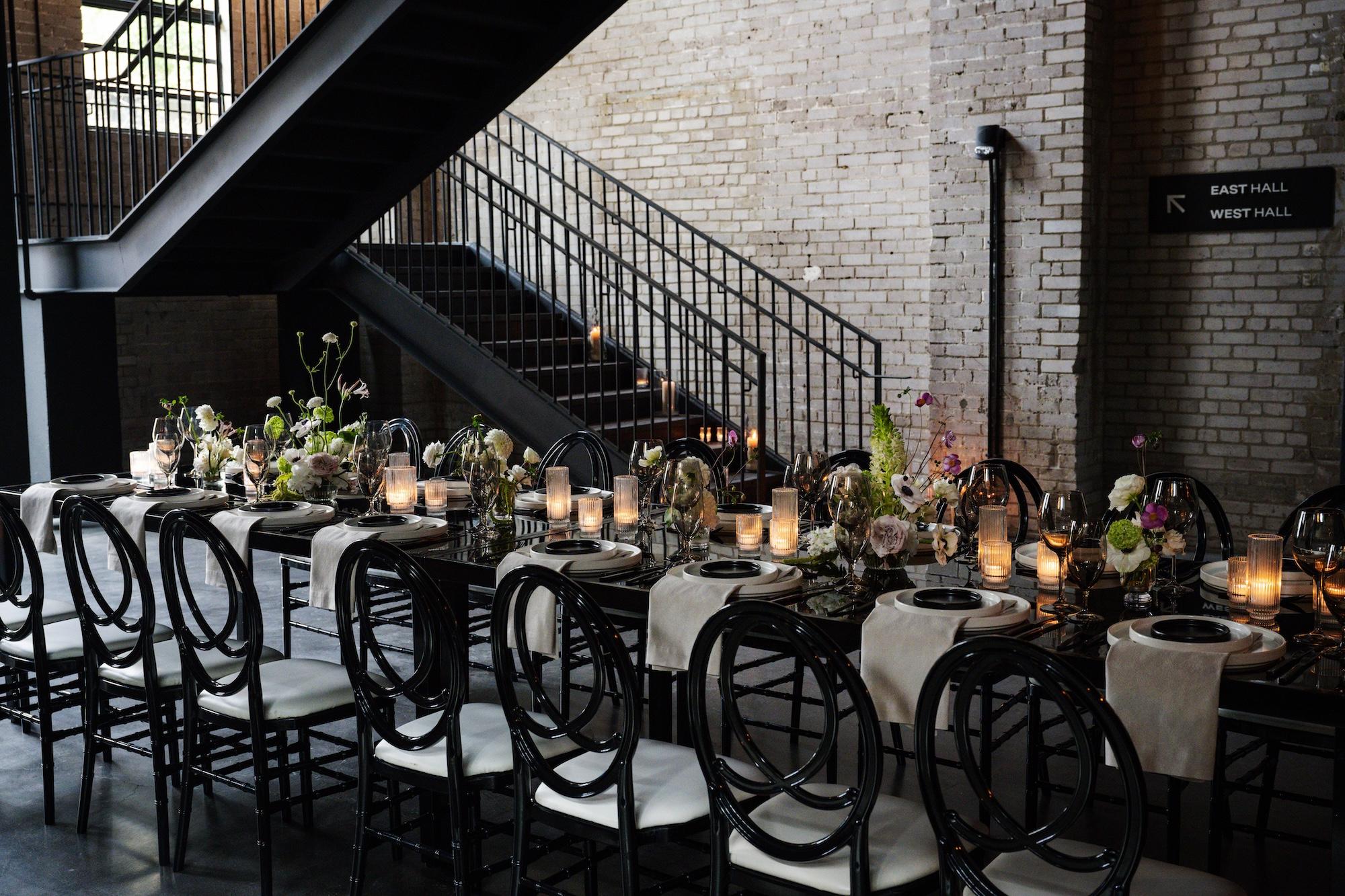 Seated dinner for 16 set in the middle of the reception of the Event Hall. The stairs leading to the West Hall are in the background and contrast against the white brick walls.