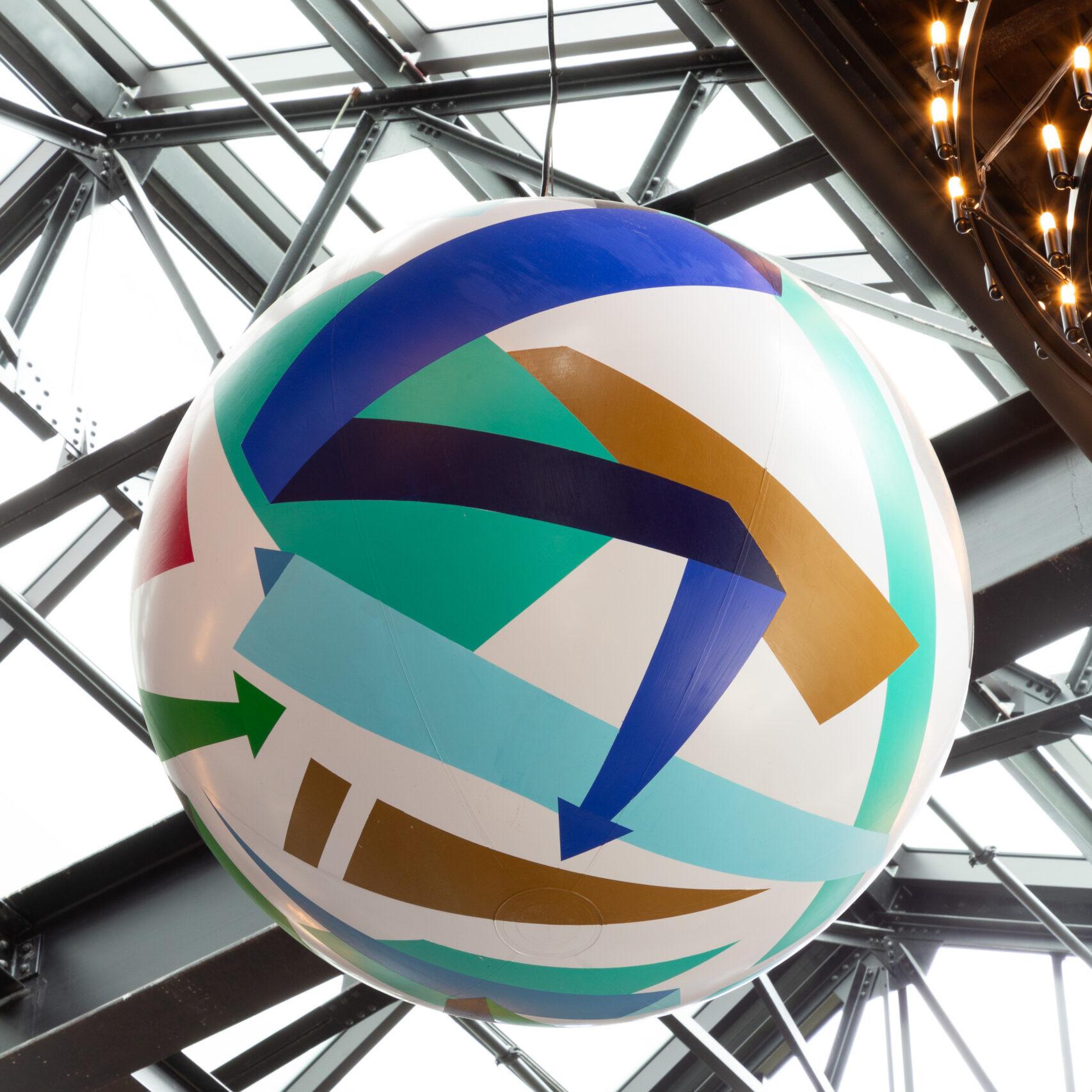 A painted sphere, depicts an array of architectural objects and geometrical elements travelling amongst one another, and hangs high above the food hall.